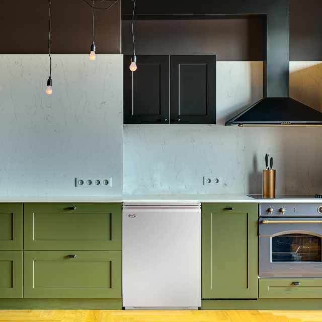 Kitchen in a modern style with a light tabletop with sink, cooker, oven, kitchen accessories. Under tabletop there are green drawers. Over tabletop there is kitchen hood, cupboard and glowing lamps.