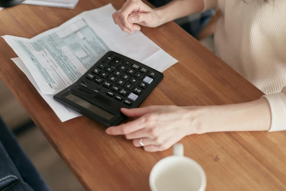 A person reviewing household bills at a wooden table while using a calculator to work out costs, illustrating how to save energy and reduce the energy bill. The scene reflects energy-efficient household choices, average electricity usage, and questions like whether radiator reflector foil works, if LED bulbs are energy-efficient, whether electricity is cheaper on weekends, and how much solar panels can save.